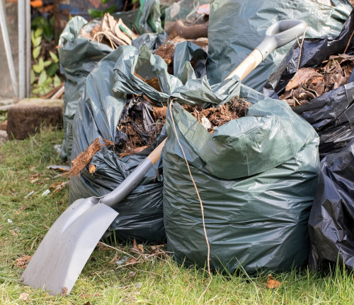 A shovel sitting amongst a pile of garbage bags that are full with garden waste.