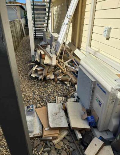 Before photo of side of house cluttered with general garden rubbish